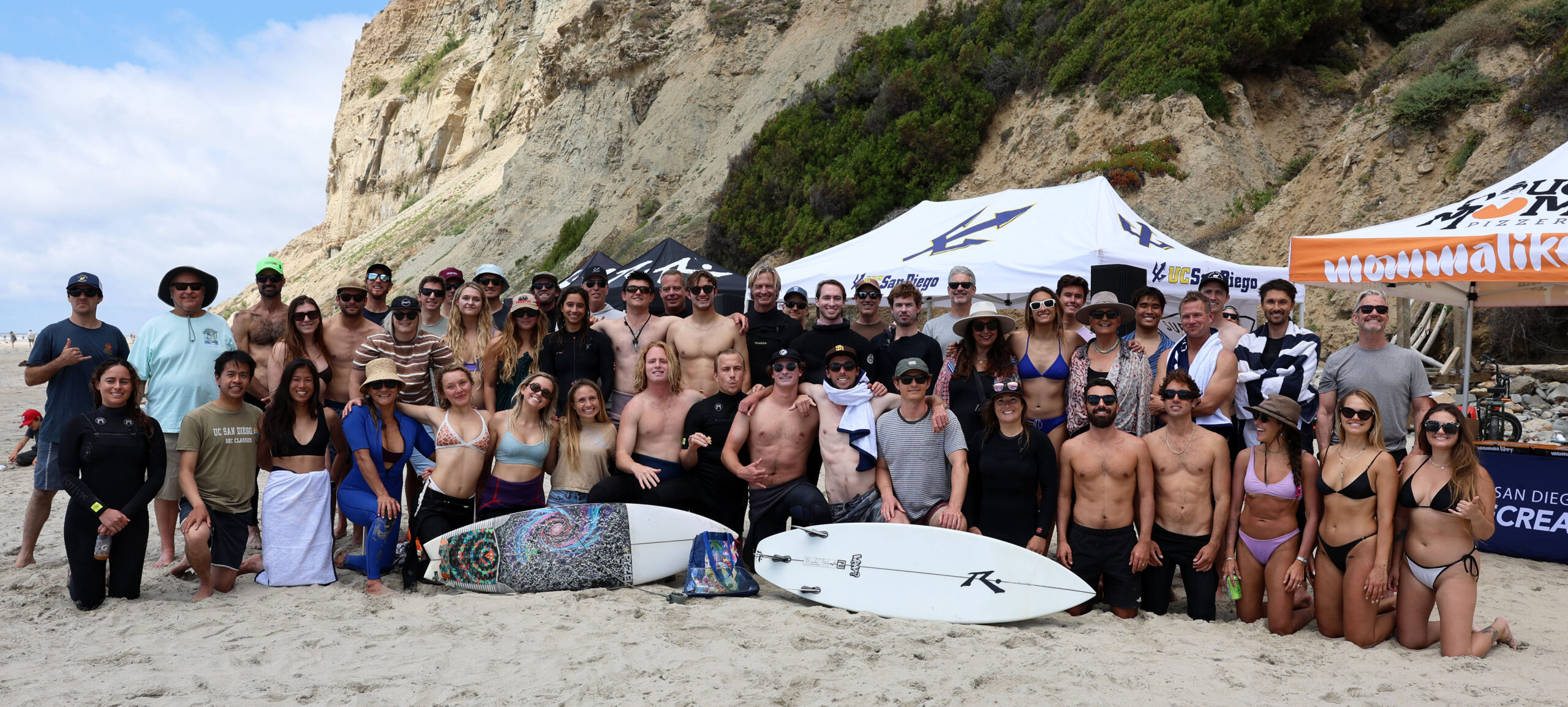 A group of male and female UC San Diego surfers poses for a photo on a beach.