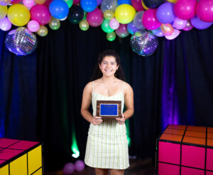 Saffron wears a green dress as she holds an award underneath colorful balloons.