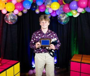 Zach is wearing a purple shirt and khakis as he holds a blue and brown award.