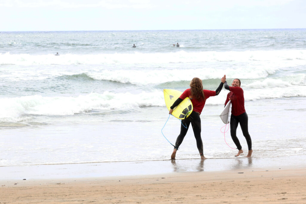 Two female surfers wearing wetsuits exchange a high-five on the beach.