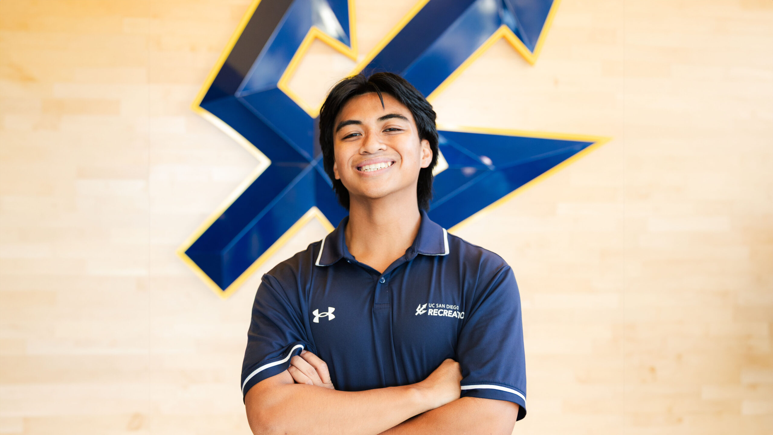 Angelo, a young man with dark hair wearing a blue polo, stands before a wooden wall with a blue UCSD Trident.
