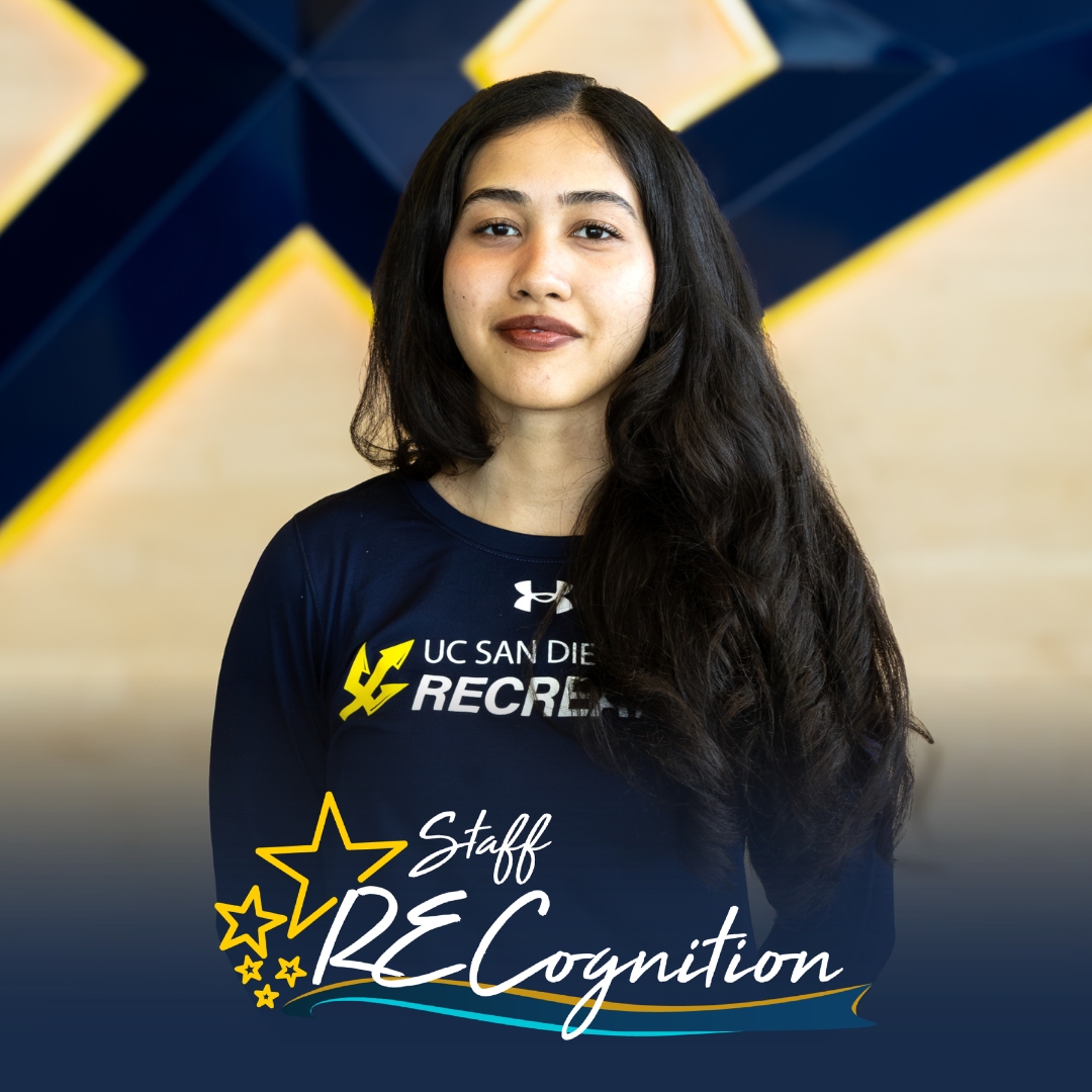 Manahil has dark hair and wears a blue longsleeve shirt with the Recreation logo. Here, she stands in front of a Trident, with the RECognition logo at the bottom.
