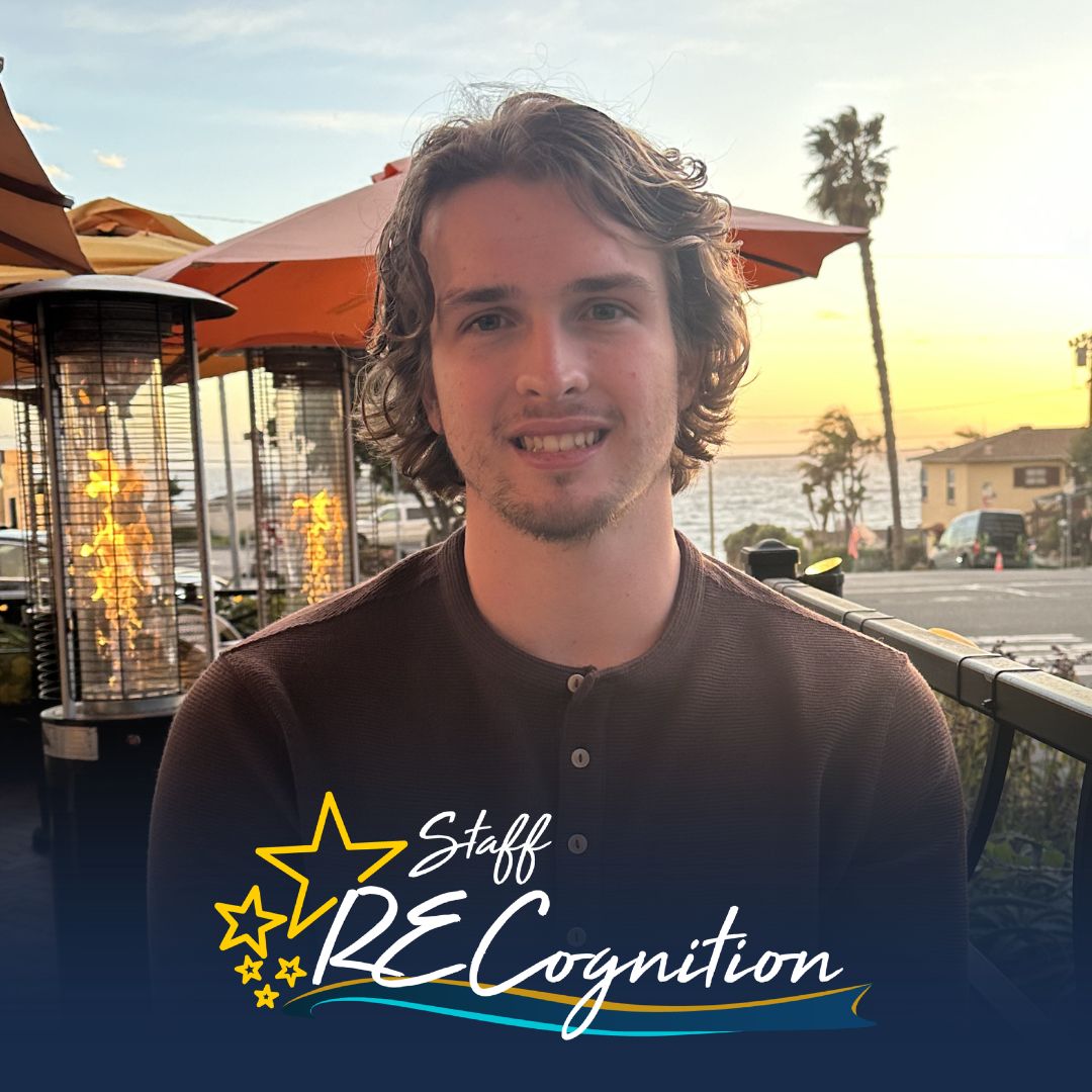 Jakob Tallman has medium length brown hair and wears a brown henley-style shirt. He sits here at a restaurant, with palm trees, umbrellas, and a sunset in the background. A Staff RECognition logo is overlayed on the bottom.