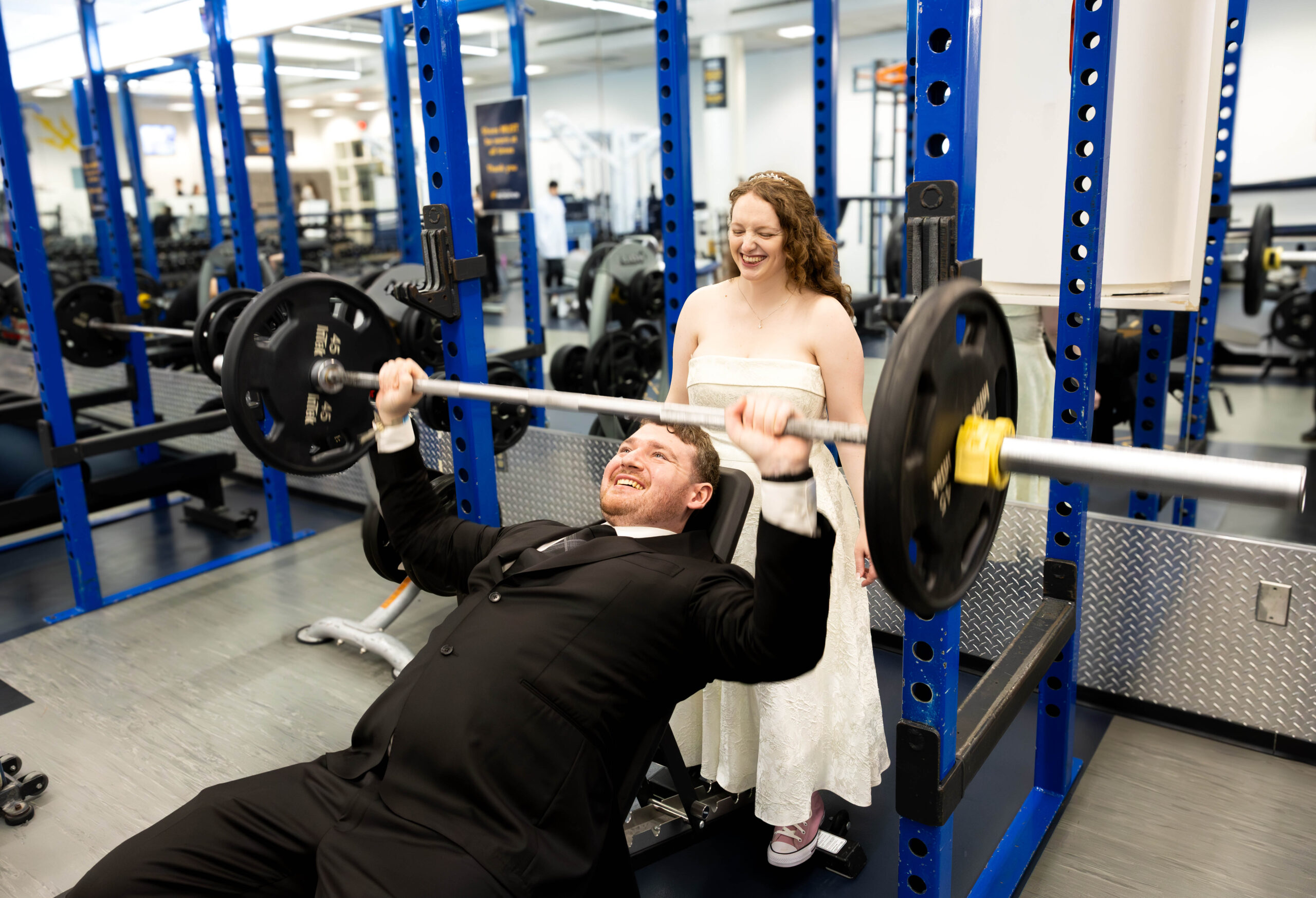 Sydney spots her husband, Paul, on an incline bench press in the UCSD weight room. Both are wearing wedding attire.
