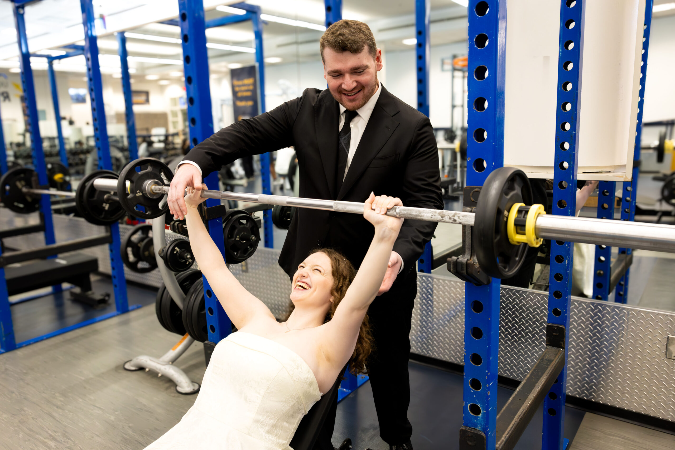 Sydney does a bench press, aided by Paul. Both are wearing wedding attire.