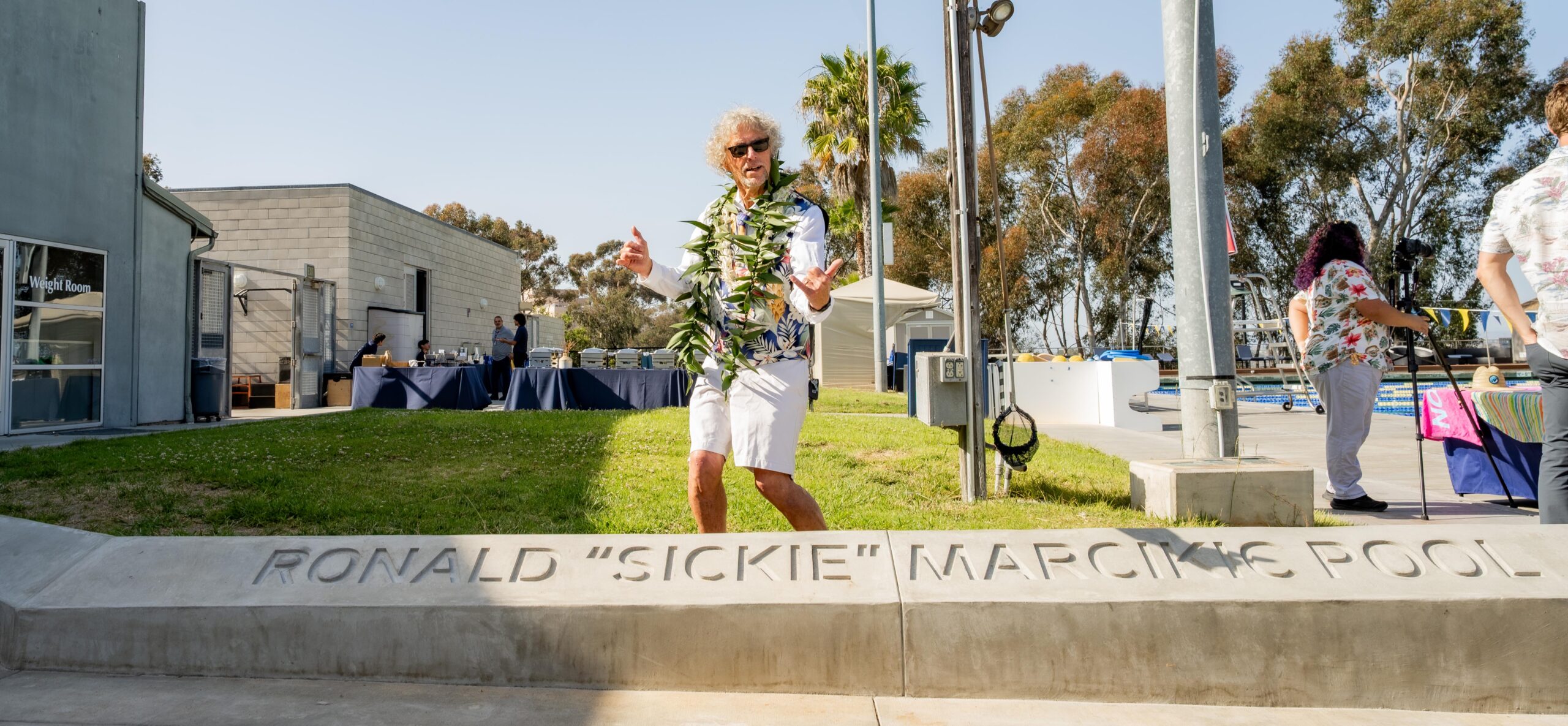 Ronald “Sickie” Marcikic, wearing a flower shirt and white shorts, stands behind a monument bearing his name. Sickie sports gray hair, sun glasses, and is giving the "shaka" sign with both hands.