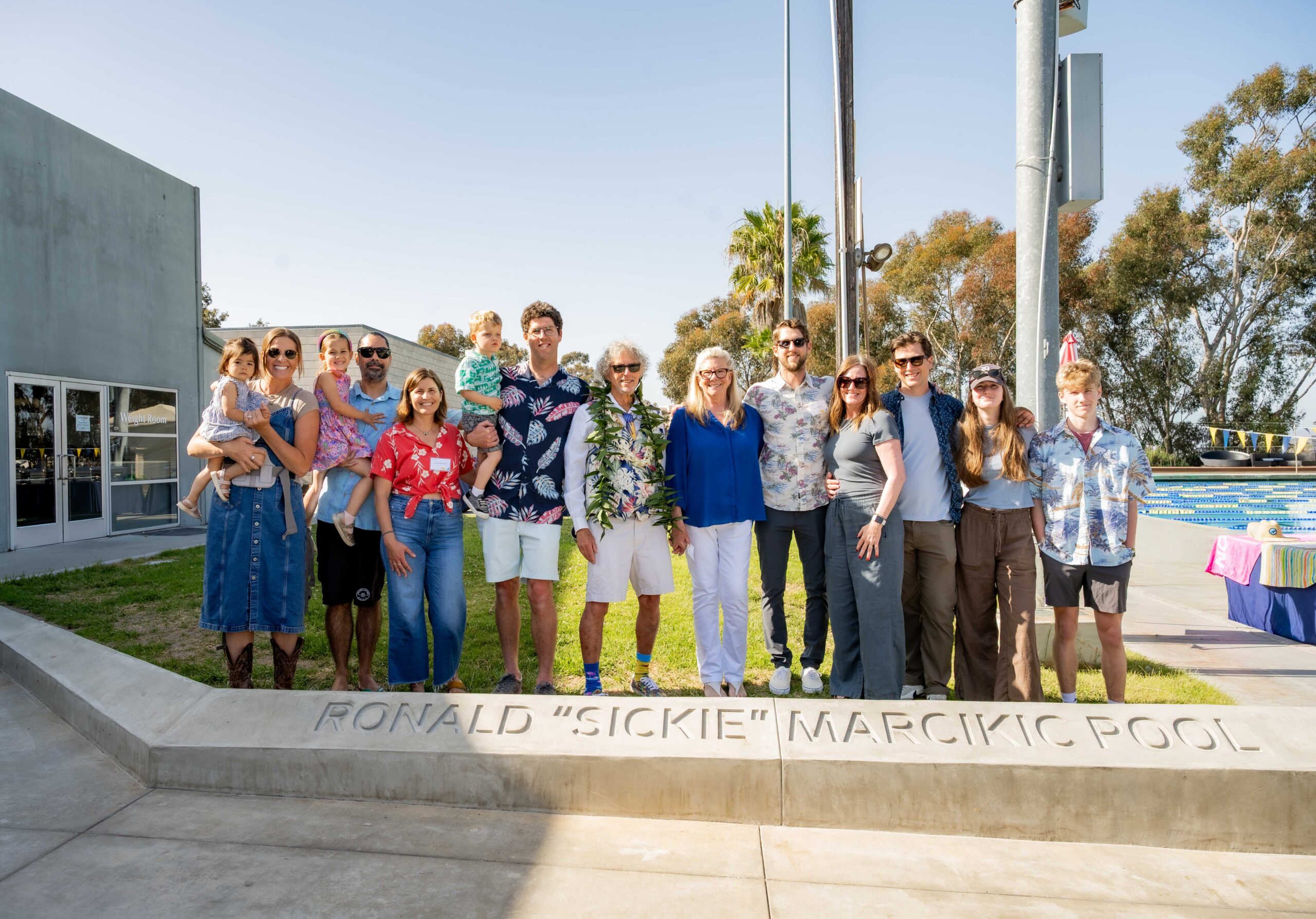 Fourteen people of all ages and genders pose for a group shot, some wearing floral shirts and sunglasses — all of them smiling.