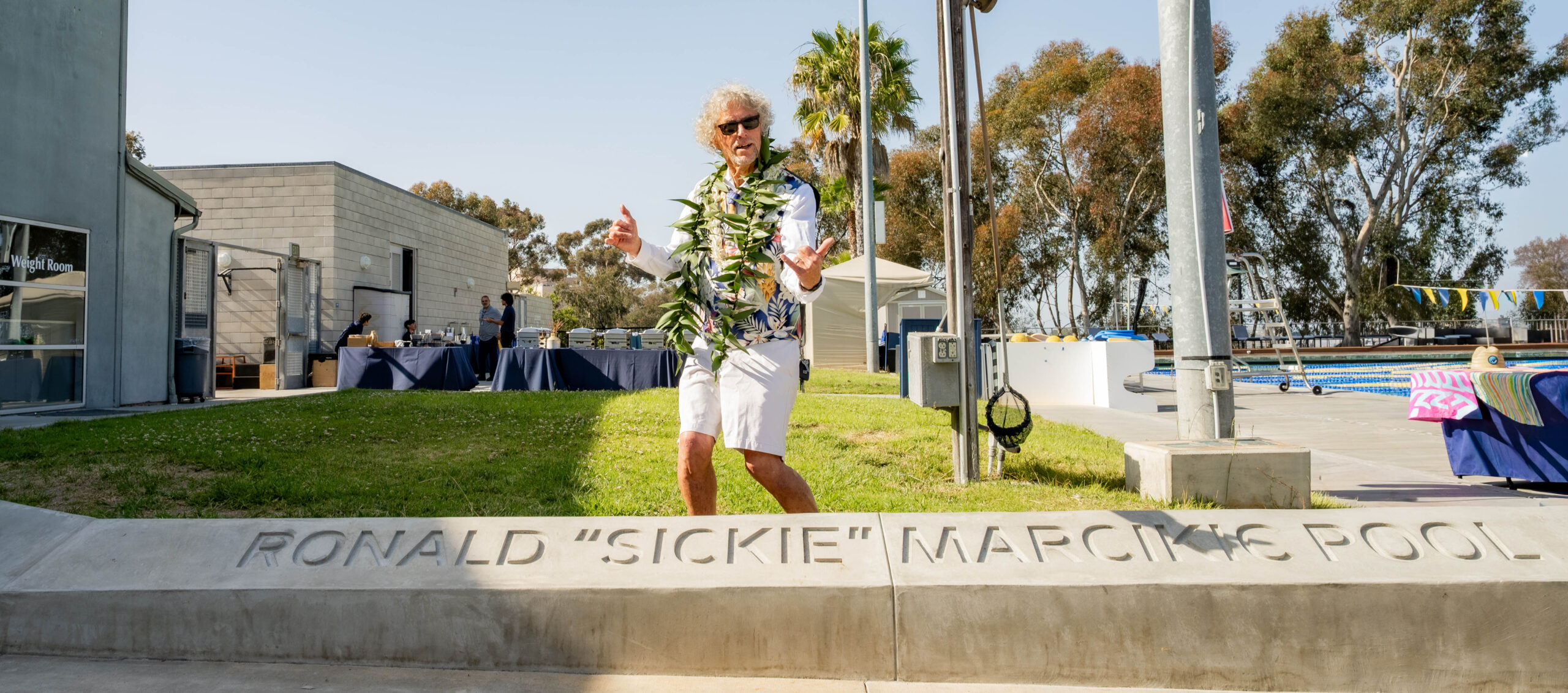 Ronald “Sickie” Marcikic, wearing a flower shirt and white shorts, stands behind a monument bearing his name. Sickie sports gray hair, sun glasses, and is giving the "shaka" sign with both hands.