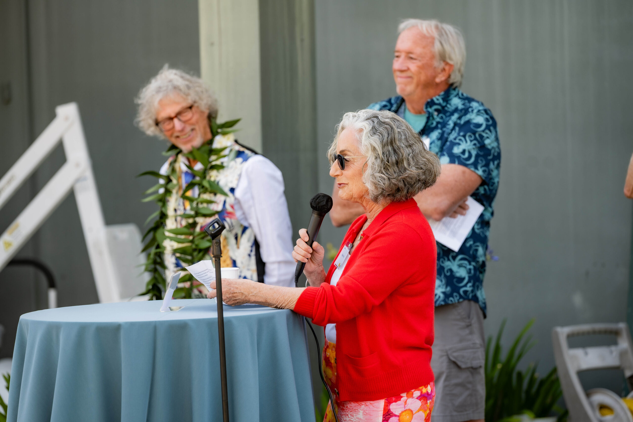 A woman in a red sweater with gray hair stands at a table to give a speech into a microphone.