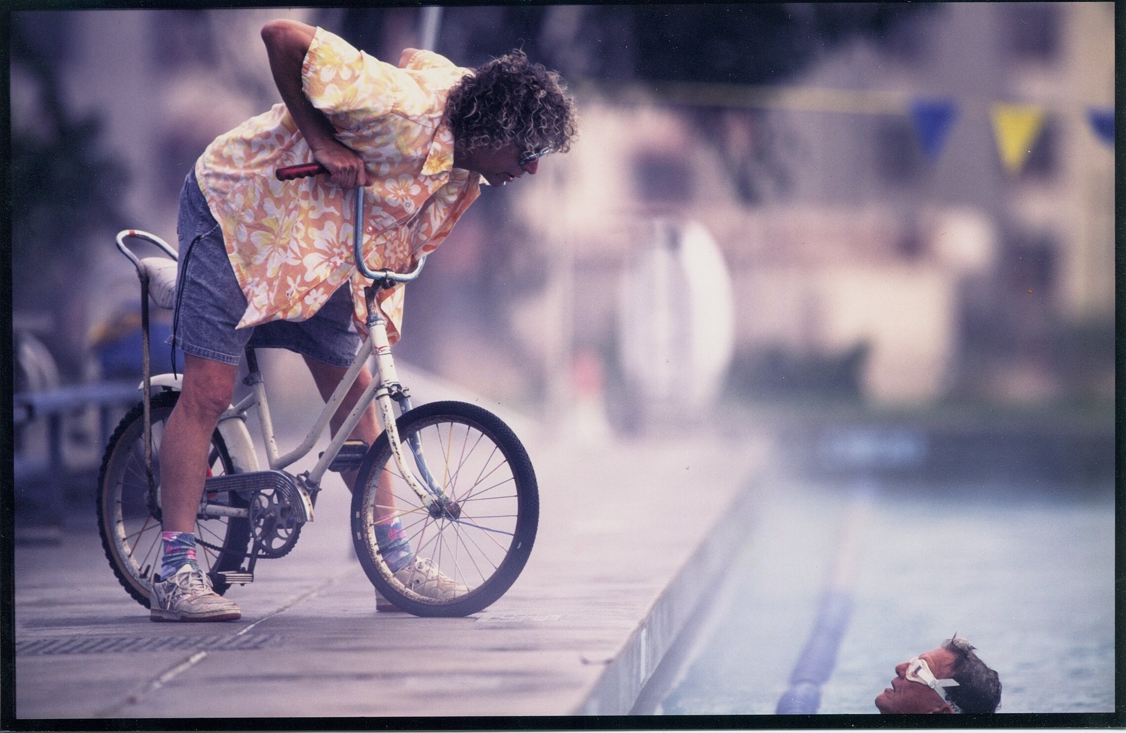 A man in a yellow shirt and shorts leans over his bicycle to talk to a swimmer in the nearby pool.